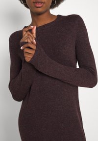 Femme portant un pull en maille à manches longues de couleur violet foncé avec des poignets côtelés, les mains jointes près de sa poitrine devant un fond clair uni.