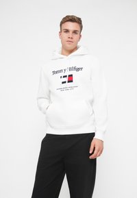 Felpa bianca con logo frontale che presenta elementi rossi, blu e bianchi. Tasca a canguro e cappuccio con stringhe. Indossata con pantaloni neri.