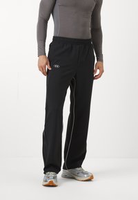 Sorte treningbukser med en elastisk midje, hvite sidestriper og et Under Armour-logo på hoften, kombinert med grå treningssko.