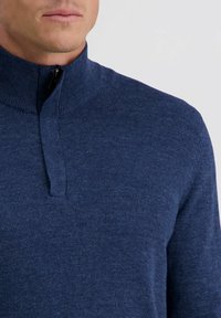 Mann in einem navyblauen Pullover mit Viertelreißverschluss und ribbeltem Kragen vor einem schlichten hellen Hintergrund.