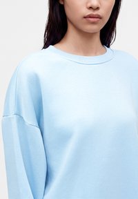 Jeune personne aux cheveux lisses et foncés portant un sweat-shirt bleu clair à col rond sur un fond uni.