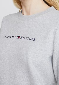 Sweatshirt gris clair avec le texte "Tommy Hilfiger" et le logo brodé rouge, blanc et bleu sur la poitrine, porté par une personne aux cheveux blonds.