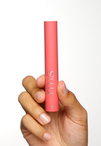 RMS läppprodukt i en cylindrisk, matt korallbehållare med silverfärgade varumärkes- och pil-detajer, hålls i en hand med neutral nagellack.