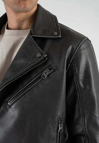 Giacca biker in pelle nera con tasche con zip, bottoni a pressione e colletto a punta. Texture liscia, vestibilità sartoriale, dettagli hardware minimi.