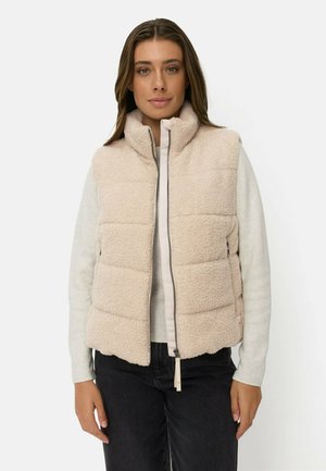 Kvinde iført beige quiltet fleecevest med lynlås over en lys langærmet top og sorte bukser, stående mod en ensfarvet hvid baggrund.