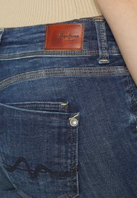 Mörkblå denimjeans med en läderetikett med "Pepe Jeans London", silverfärgade detaljer och en distinkt vågig sömdekoration på fickan.