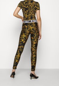 Schwarz-goldene leggings mit Muster und einem passenden kurzärmligen Oberteil, das einen weißen, mit Logo versehenen Bund und eine figurschmeichelnde Passform aufweist.