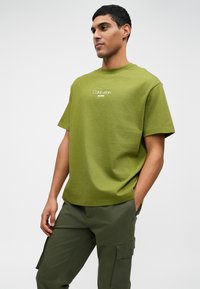 Olive zelena pamučna majica kratkih rukava s okruglim izrezom. Sadrži bijeli natpis "Calvin Klein Jeans" na prsima.