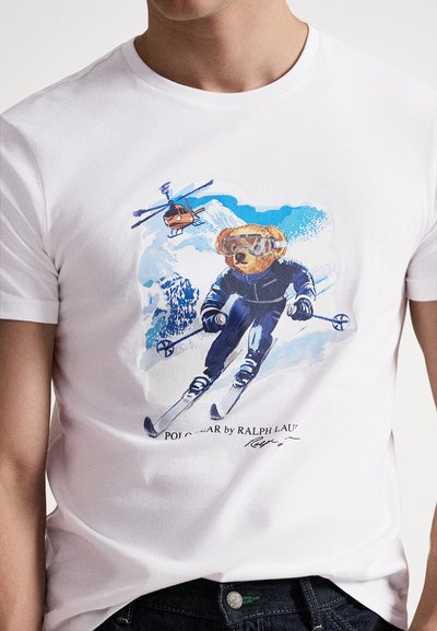 Bijela pamučna majica s printom medvjeda koji skija u naočalama, uz helikopter i plavu planinsku pozadinu.