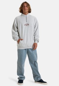 Šedá fleece mikina na půlzip s volným střihem, s vyšitým logem na hrudi, kombinovaná s světle modrými volnými kalhotami a teniskami.
