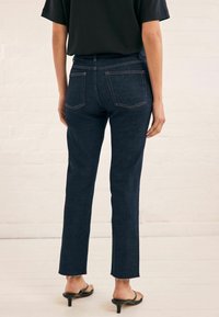 Jeans de mezclilla azul oscuro con corte recto, dobladillo deshilachado y dos bolsillos traseros. Combinados con una blusa negra y sandalias de tacón.