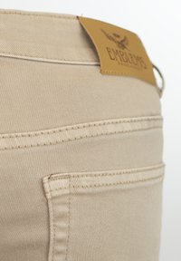 Jeans de denim beige con una textura suave, que presentan un parche de cuero con el logotipo y costuras detalladas cerca de la cinturilla y los bolsillos.