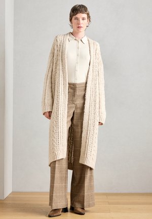 Cardigan beige oversized in maglia con motivo testurizzato, abbinato a una camicia bianca con bottoni e pantaloni a vita alta a quadri marroni. Stivali con tacco marroni.