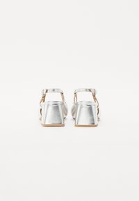 Silver heeled sandals with a metallic finish, an ankle strap, and a triangular-shaped chunky heel. Soft footbed and smooth texture.