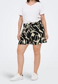 Mujer vestida con camiseta blanca de cuello en V, pantalones cortos con estampado abstracto en negro y beige con cordón, y deportivas blancas, de pie frente a un fondo liso.