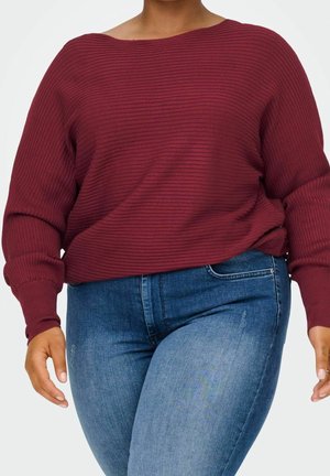 Personne portant un pull côtelé bordeaux avec des manches bouffantes et un jean skinny taille haute bleu sur un fond uni.