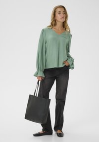 Maglietta verde a maniche lunghe con scollo a V e polsini arricciati, abbinata a jeans scuri e una borsa a tracolla nera. Scarpe nere flat completano il look.