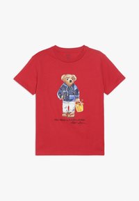 Camiseta de algodón roja con un gráfico de un oso con chaqueta azul y pantalones blancos, sosteniendo un cubo amarillo. El texto dice "POLO BEAR by RALPH LAUREN."