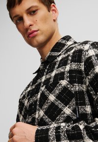 Camisa a cuadros en blanco y negro con tejido texturizado, cuello abotonado y mangas largas con botones negros y detalle de etiqueta de logo.