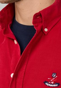 Rotes Cordhemd mit Button-Down-Kragen, ausgestattet mit weißen Knöpfen und einem dunkelblauen T-Shirt darunter, das ein gesticktes Logo zeigt.