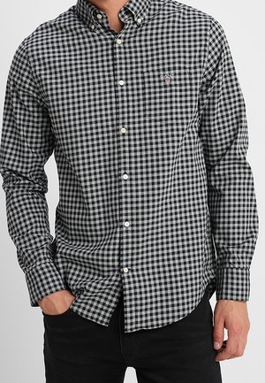 Man in een zwart-wit geruite blouse met lange mouwen en zwarte broek, met één hand in zijn zak.