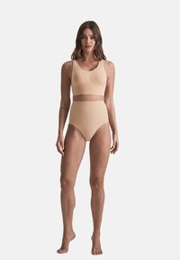 Donna in piedi a piedi nudi, che indossa un reggiseno a vita alta beige e un top corto senza maniche abbinato, su uno sfondo bianco semplice.