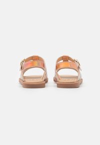 Sandalias planas marrones con dos tiras iridiscentes brillantes y una hebilla lateral. La suela es plana y está hecha de goma marrón clara.