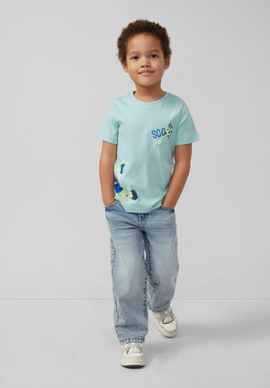 Jonge jongen staand met handen in zakken, dragen een lichtblauw voetbalthema T-shirt, losse blauwe jeans en witte sneakers tegen een effen achtergrond.