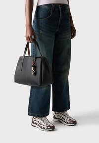 Borsa in pelle nera con forma strutturata, accentuata da un portachiavi decorativo, abbinata a un paio di jeans in denim blu larghi e sneakers con motivo.