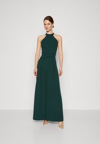 Grønn halterneck jumpsuit med en tettsittende bodice, plissert vid benbukse og glatt tekstur. Tilbehørt med dekorative hæler.