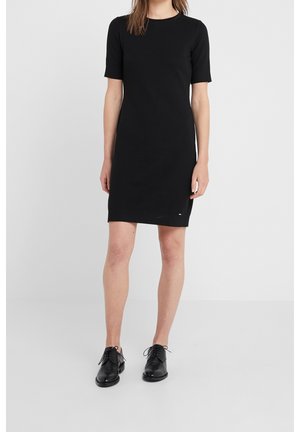 Kobieta ubrana w czarną sukienkę z krótkim rękawem do kolan oraz czarne, sznurowane buty, stojąca na jednolitym jasnym tle.