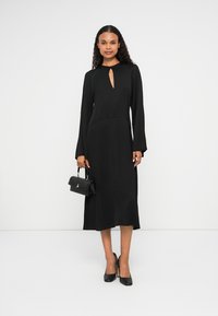 Robe noire à manches longues avec un décolleté en trou de serrure, texture lisse et silhouette ajustée. Associée à des escarpins noirs et un petit sac à main.