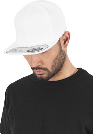Căciulă albă ajustată cu vârf plat, având orificii de ventilație și un eticheta pe partea inferioară a vârfului, purtată peste un tricou negru.