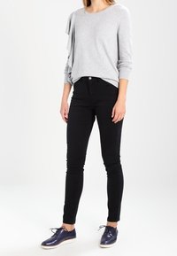 Grauer Pullover mit einem Rüschen-Detail an einem Ärmel, kombiniert mit schwarzen Slim-Fit-Jeans und glänzenden marineblauen Schuhen. Glatte Textur und lässiges Design.