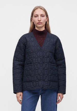 Femme aux longs cheveux blonds porte une veste matelassée bleu marine sur un pull à col roulé bordeaux et un jean bleu, debout devant un fond uni.