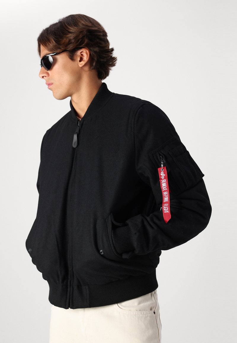 Schwarze Bomberjacke mit gerippten Bündchen, Reißverschluss, Seitentaschen und einem roten Etikett am Ärmel. Hergestellt aus einem glatten, strapazierfähigen Stoff.