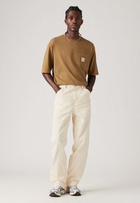 Camiseta marrón de manga corta con bolsillo en el pecho, combinada con pantalones sueltos color crema. Los accesorios incluyen una cadena de plata y zapatillas.