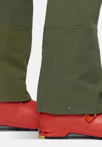 Bottes de ski rouges avec une finition texturée, associées à un pantalon vert olive présentant une section inférieure à motif et un matériel argenté à l'ourlet.