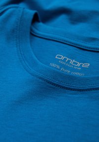 T-shirt di cotone blu brillante con scollatura rotonda, caratterizzato da una consistenza liscia e un'etichetta che riporta "linea di cotone ombre 100% cotone puro."