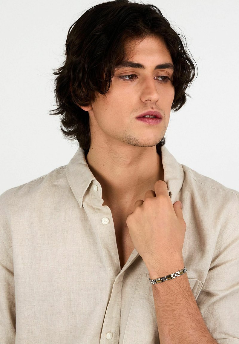 Bracciale d'argento con maglie intrecciate dalla texture lucida, indossato su una mano contro una camicia di lino chiaro.