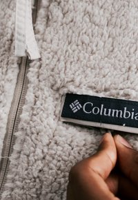 Mano che tiene una giacca in pile beige vicino all'etichetta del marchio Columbia e alla cerniera.