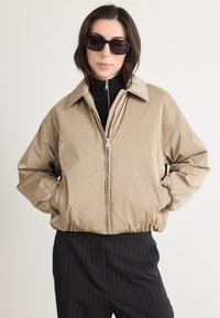 Beige, lightweight jacket with a collar, front zipper, and elastic cuffs. Worn over a black turtleneck and paired with pinstriped pants.