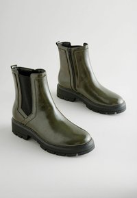 Botas de tobillo de cuero verde con un acabado brillante, paneles laterales elásticos, cierre de cremallera y suelas de goma negras. Textura suave y diseño moderno.