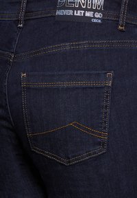 Jeans de mezclilla de color azul oscuro con costuras naranjas, un bolsillo trasero con un diseño sutil y una etiqueta de cuero de marca en la cintura.