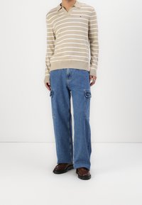 Polo en maille rayée beige et blanche avec un col en V ; associé à un jean cargo large bleu clair avec des poches avant ; chaussures marron.