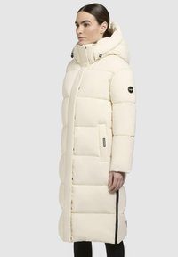 Long, puffy coat in light cream color, featuring a hood, zippered pockets, and a quilted design. Made with smooth, soft fabric.