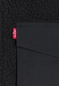 Tissu noir texturé avec un patch rectangulaire noir cousu et une petite étiquette rouge Levi's sur le bord gauche.