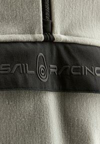 Lekka szara odzież polarowa z czarnym panelem materiału. Posiada zamek błyskawiczny i wypukłe czarne logo "SAIL RACING" na klatce piersiowej.