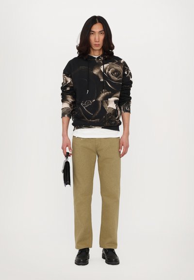 Svart hoodie med blommönster, tillsammans med ljus khaki-byxor. Modellen håller en svart väska och bär svarta skor.
