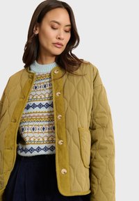 Olijfgroene gesteepte jas met een corduroy kraag en knopen, gedragen over een gebreide trui met een patroon in blauw-, geel- en crèmekleuren.